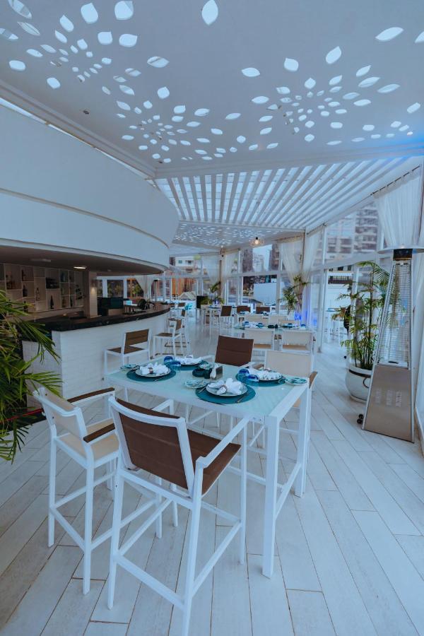 Sunrise Alex Avenue Hotel Alexandria Eksteriør bilde The photo shows a bright and modern restaurant interior. The space features white and light-colored furniture, including tables with a combination of white and brown chairs. Each table is elegantly set with plates, utensils, and decorative elements. 