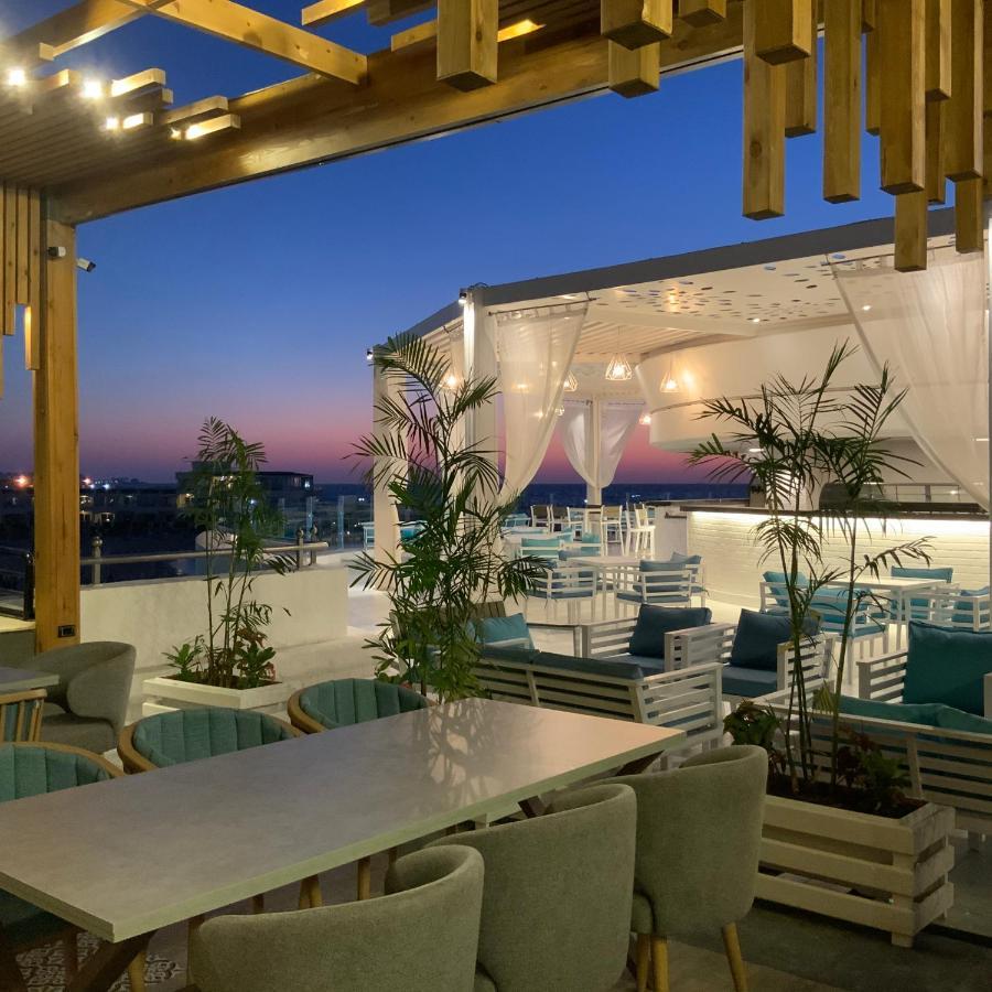 Sunrise Alex Avenue Hotel Alexandria Eksteriør bilde The photo shows a stylish outdoor dining area or terrace set against a beautiful sunset. There are several tables and chairs arranged neatly under a covered structure, with a backdrop of soft white curtains. The space is decorated with potted plants,
