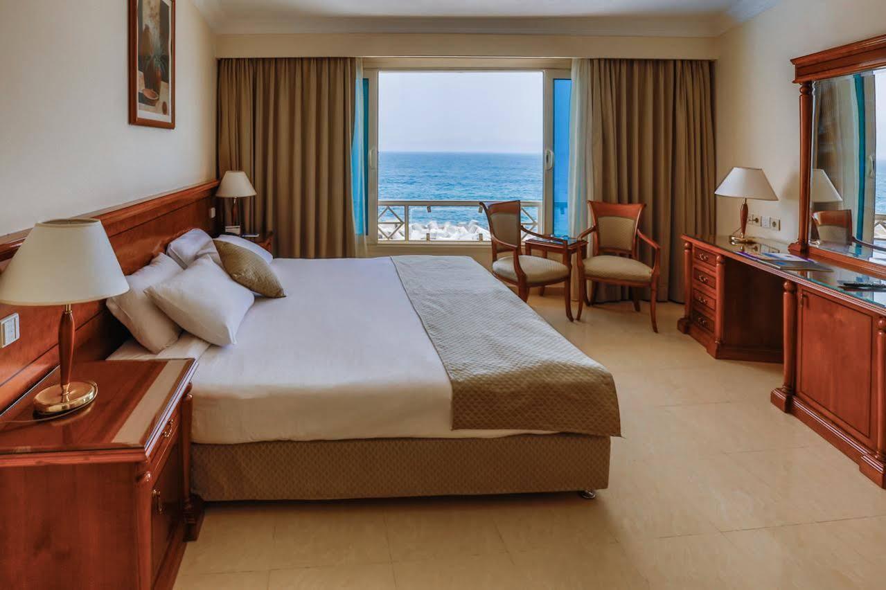 Sunrise Alex Avenue Hotel Alexandria Eksteriør bilde The photo shows a hotel room featuring a large bed with white linens and decorative pillows. There are two wooden chairs positioned near a window, which offers a view of the ocean. The room has beige walls and light-colored curtains that frame the wi