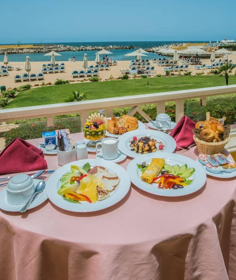 Sunrise Alex Avenue Hotel Alexandria Eksteriør bilde The photo shows a beautifully set breakfast table on a balcony with a scenic view of a beach. There are several plates of food arranged attractively. One plate features an assortment of fruits, including slices of melon, berries, and other colorful f