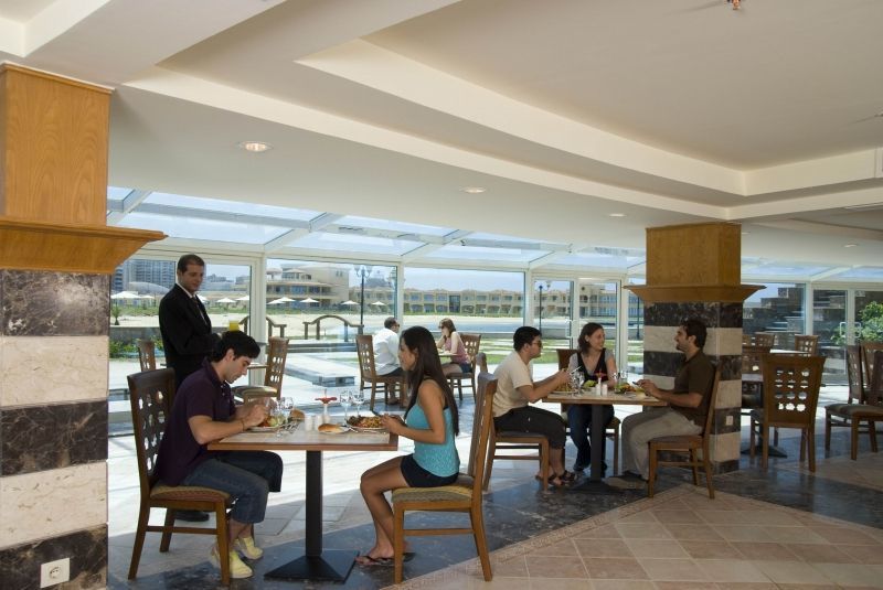 Sunrise Alex Avenue Hotel Alexandria Restaurant bilde The photo shows an indoor dining area, possibly in a restaurant or café. There are several tables with people sitting at them. Two groups of diners are engaged in conversation while enjoying their meals. A server is also present, attending to one of 
