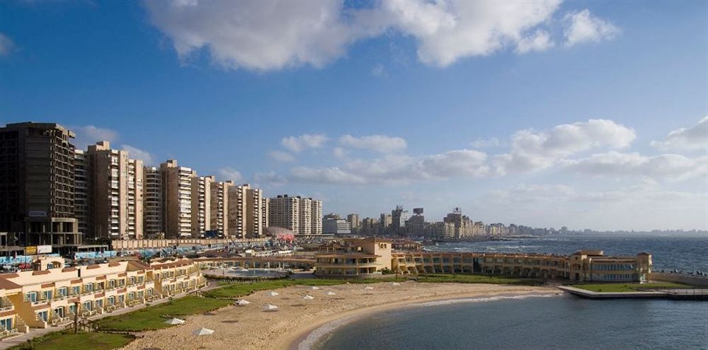 Sunrise Alex Avenue Hotel Alexandria Eksteriør bilde The photo shows a scenic coastal view featuring a sandy beach and a curved shoreline. In the foreground, there are several sun loungers and a landscaped area. Beyond the beach, there are multiple high-rise buildings lining the waterfront, indicating 
