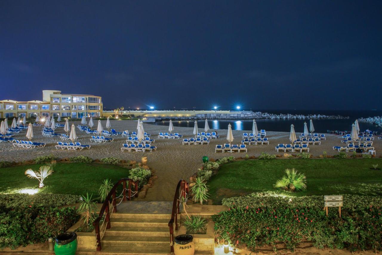 Sunrise Alex Avenue Hotel Alexandria Eksteriør bilde The photo shows a beach scene at night. In the foreground, there are steps leading down from a grassy area lined with plants. The beach is filled with numerous lounge chairs, all arranged with umbrellas, suggesting a relaxed atmosphere. In the backgr