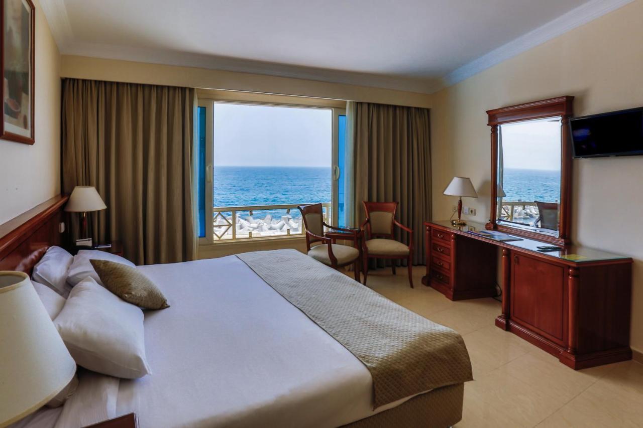 Sunrise Alex Avenue Hotel Alexandria Eksteriør bilde The photo shows a hotel room with an ocean view. The room features a large bed with neutral-colored bedding and pillows. There are curtains covering the windows, allowing plenty of light to enter. Beside the bed, there is a wooden desk and a chair po