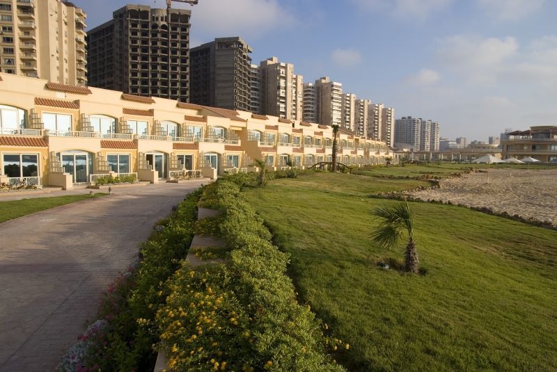 Sunrise Alex Avenue Hotel Alexandria Eksteriør bilde The photo depicts a scenic view of a shoreline area with modern buildings in the background. In the foreground, there is a well-maintained green lawn, bordered by a pathway. The buildings appear to be multi-story residential or commercial structures,