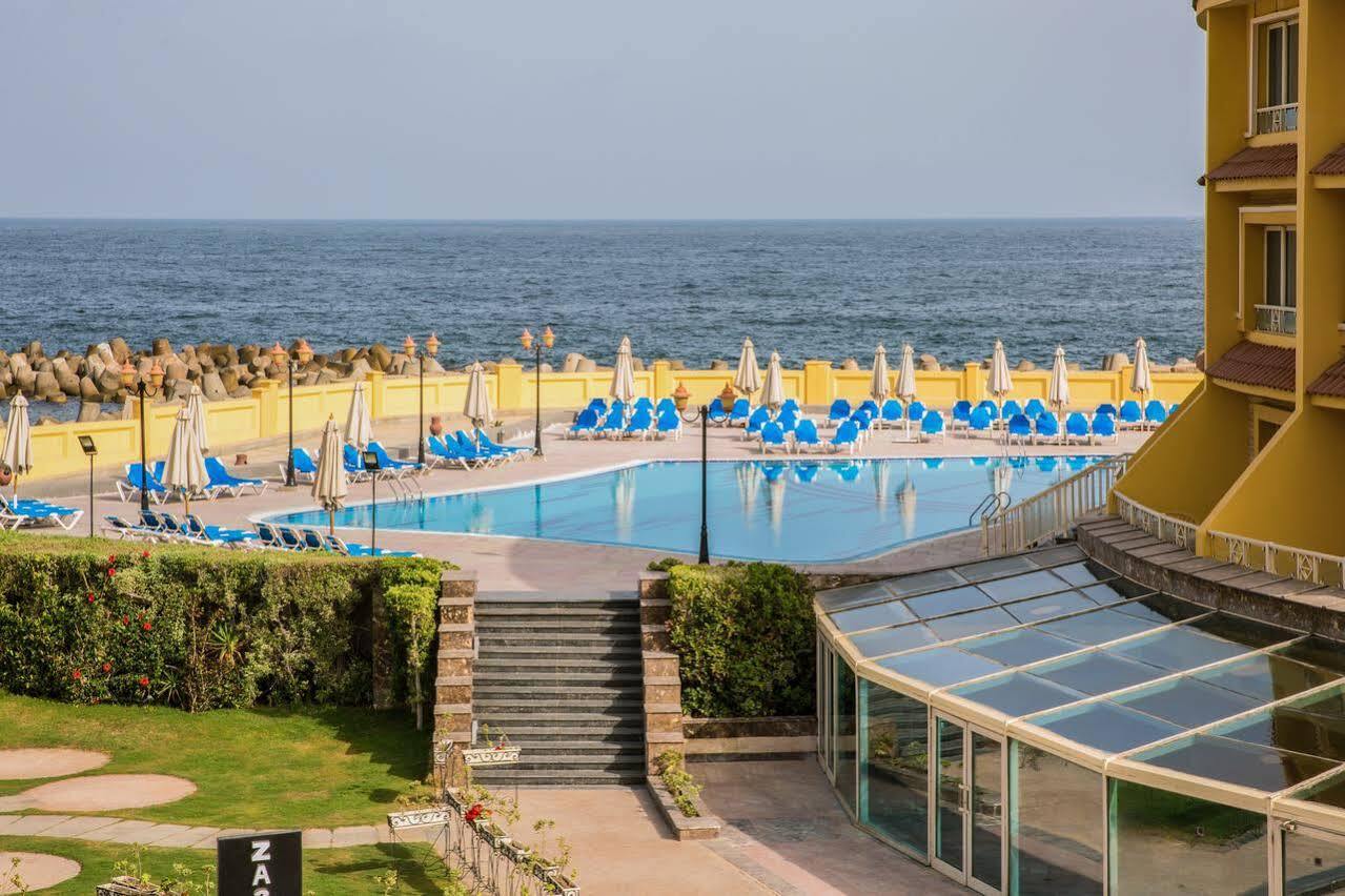 Sunrise Alex Avenue Hotel Alexandria Eksteriør bilde The photo depicts a scenic view of a pool area adjacent to the ocean. In the foreground, there is a large swimming pool surrounded by numerous blue lounge chairs and umbrellas, indicating a relaxing setting. There are steps leading down from a grassy