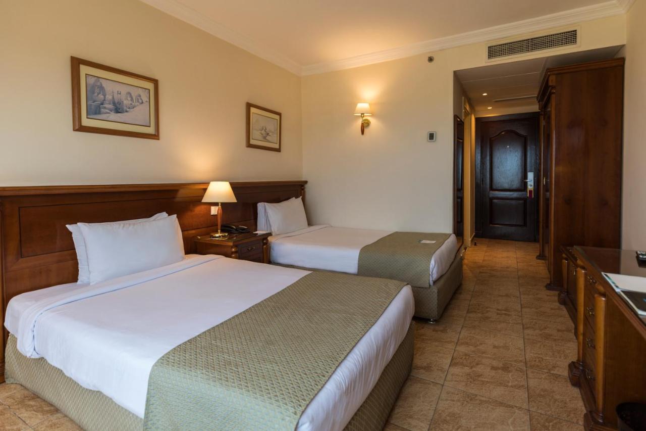 Sunrise Alex Avenue Hotel Alexandria Rom bilde The photo shows a hotel room featuring two twin beds with light-colored bed linens and green decorative blankets. There's a bedside table with a lamp next to one of the beds. The walls are painted a light color, and there are two framed pictures hang