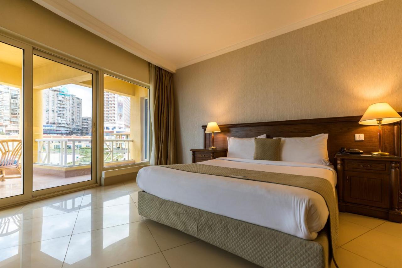 Sunrise Alex Avenue Hotel Alexandria Rom bilde The photo shows a spacious hotel room featuring a large bed with white linens and decorative pillows. There are wooden nightstands with lamps on both sides of the bed. The walls are painted in neutral tones, and the floor is tiled. Large windows prov